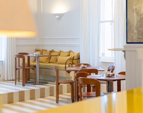 Bright hotel dining area with wooden chairs, round tables, cushioned bench seating, white curtains, and soft wall lighting