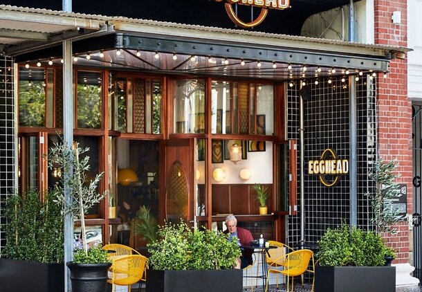 Cozy outdoor seating at Egghead café with yellow chairs, greenery in planters, string lights, and wood-framed windows