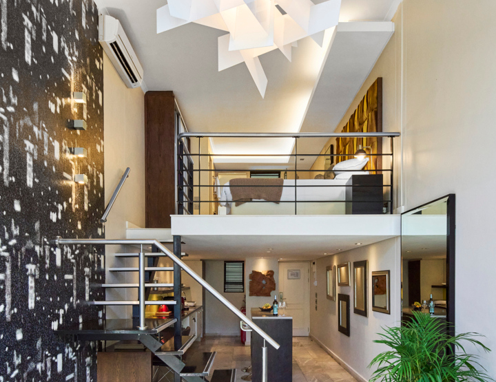 Modern loft suite with mezzanine bedroom, contemporary staircase, abstract pendant light, sleek kitchenette, and stylish living area with plants.