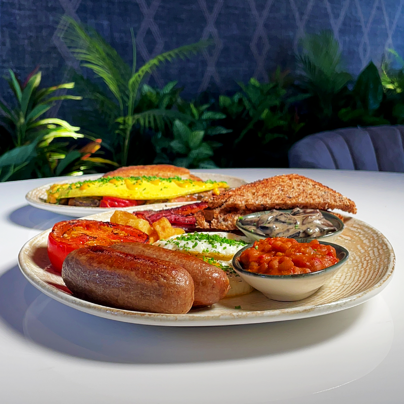 Hearty breakfast plate with sausages, grilled tomato, sautéed mushrooms, beans, toast, and an omelette, served in a stylish dining setting