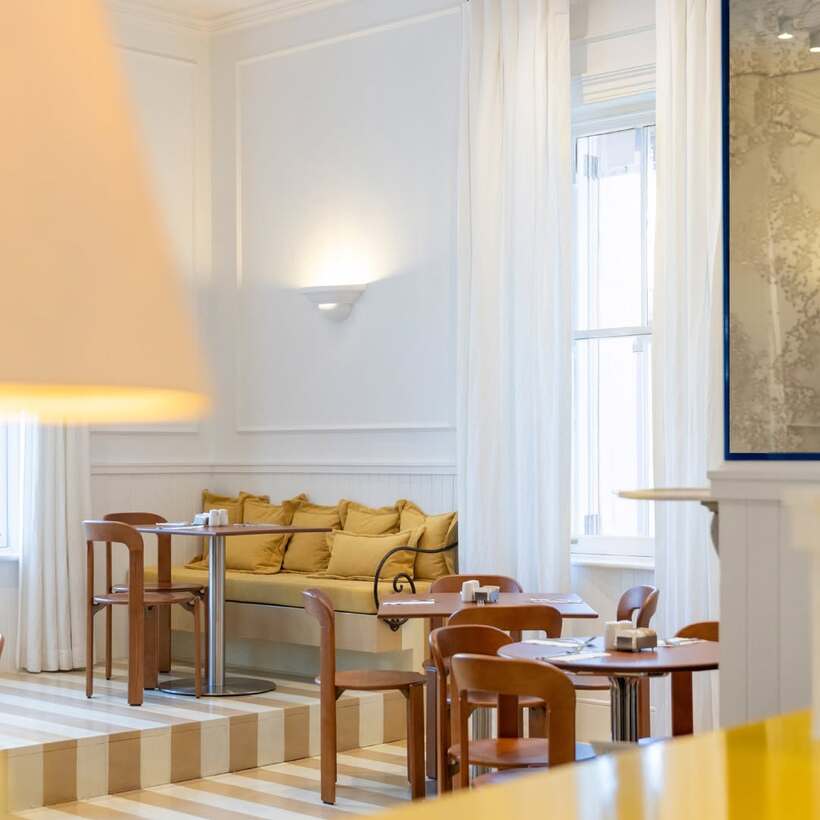 Bright hotel dining area with wooden chairs, round tables, cushioned bench seating, white curtains, and soft wall lighting