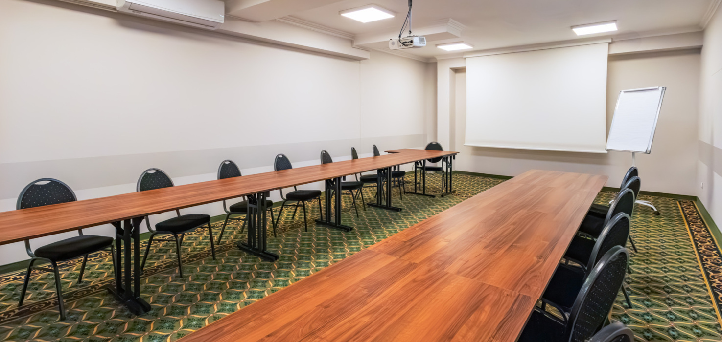 Sala konferencyjna z długimi stołami, krzesłami, projektorem i flipchartem, idealna na spotkania biznesowe.