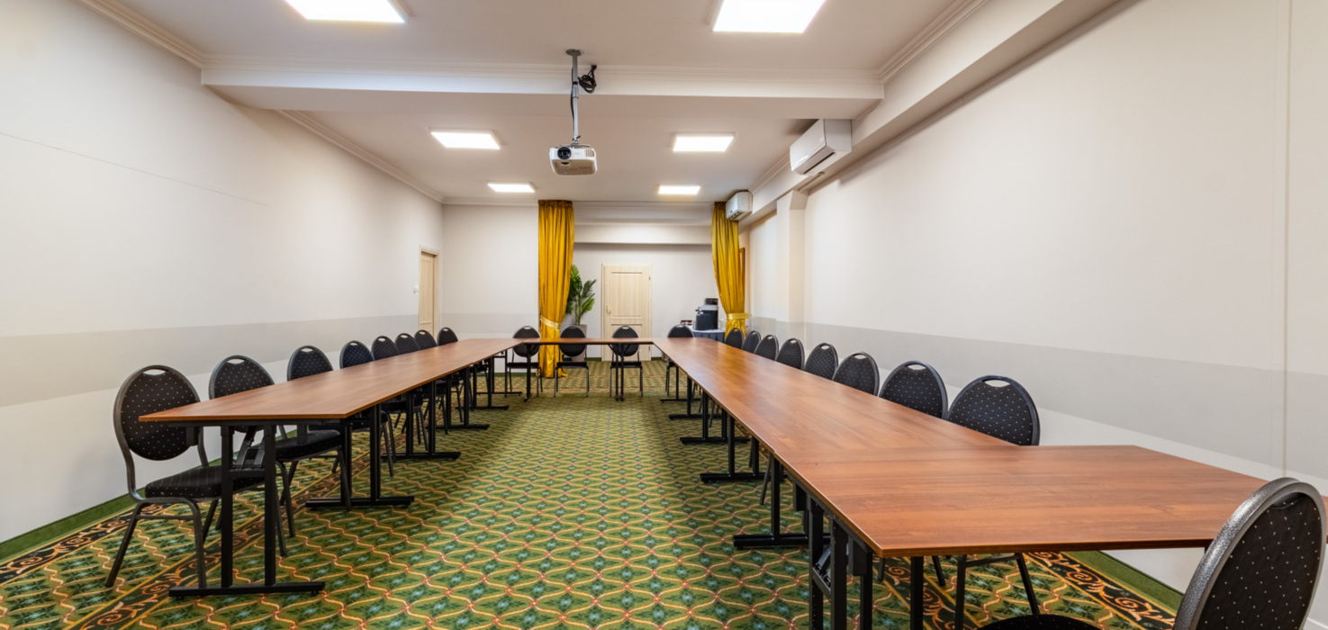 Konferencyjna sala z długim stołem w kształcie litery U, wygodnymi krzesłami i projektorem pod sufitem.