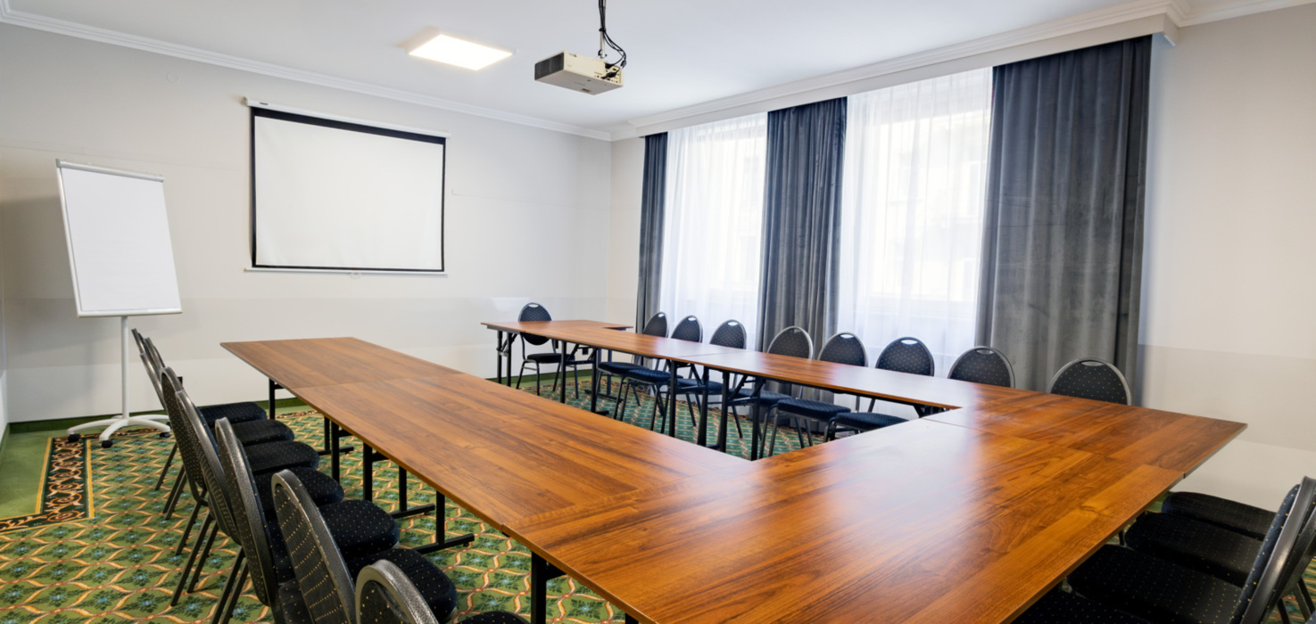 Modern conference room with U-shaped wooden table, chairs, ceiling-mounted projector, and projection screen on the wall.