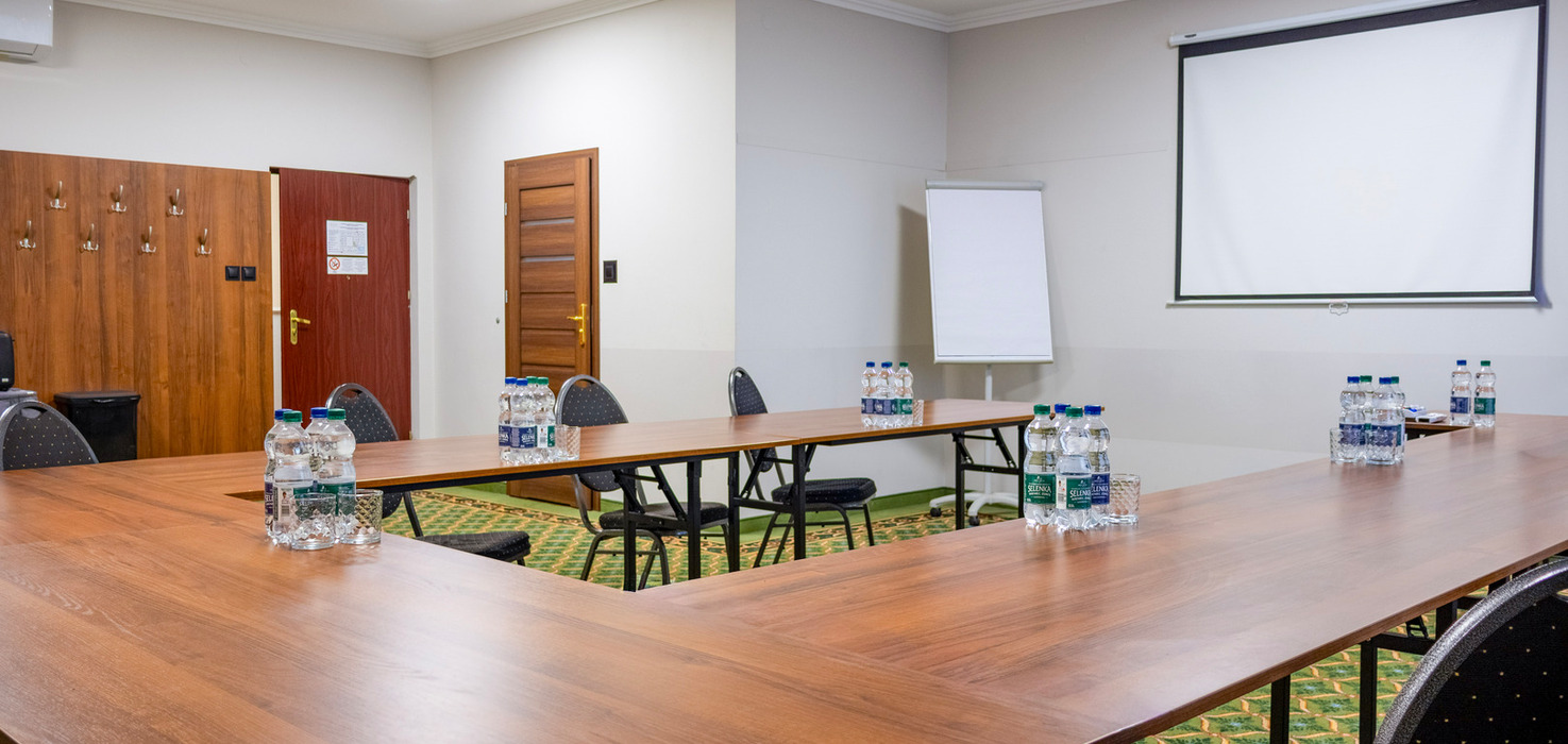 Sala konferencyjna w układzie U z projektorem, ekranem, flipchartem i butelkowaną wodą na ozdobnym dywanie.