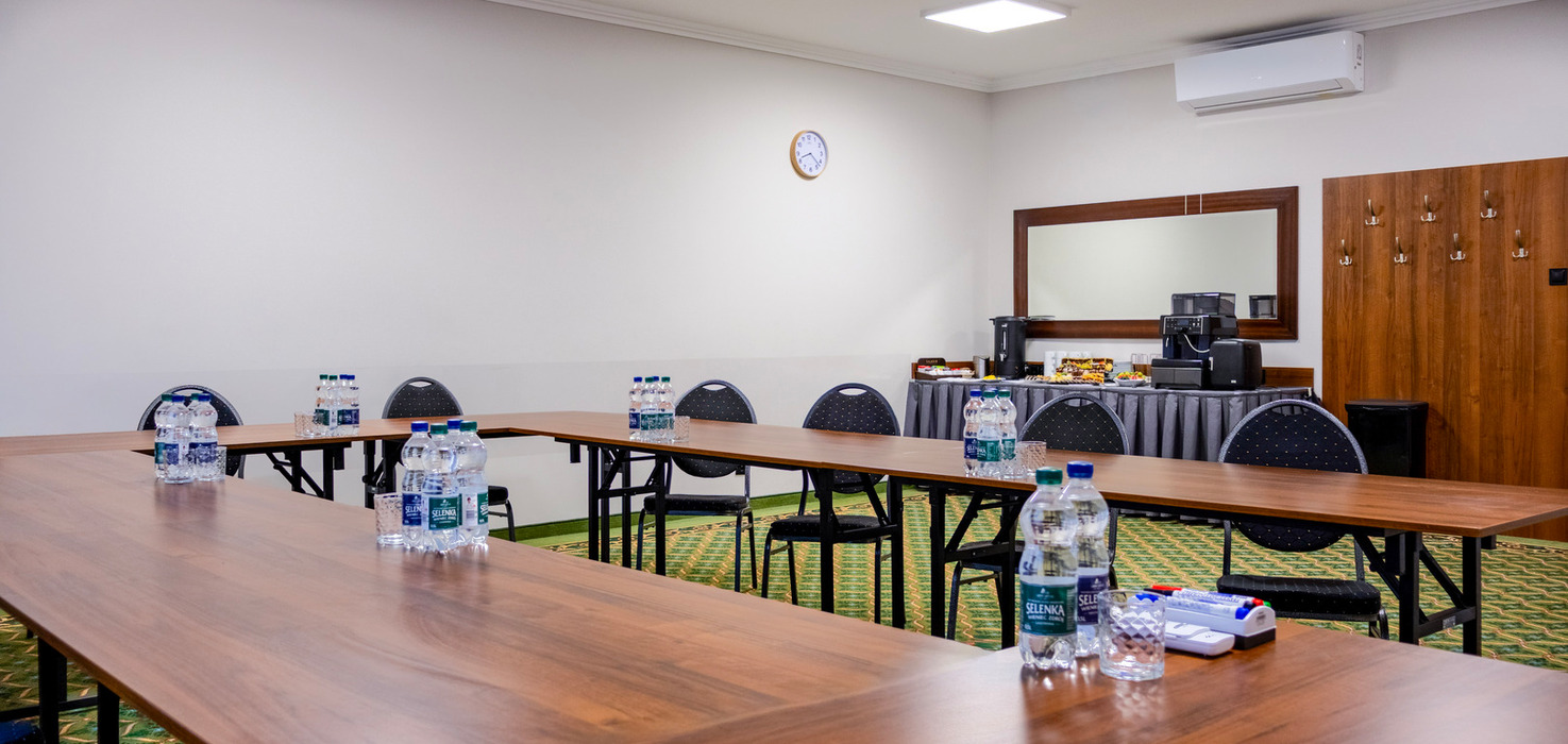 Sala konferencyjna w układzie U z drewnianymi stołami, butelkowaną wodą, projektorem sufitowym, stacją kawową i klimatyzacją.