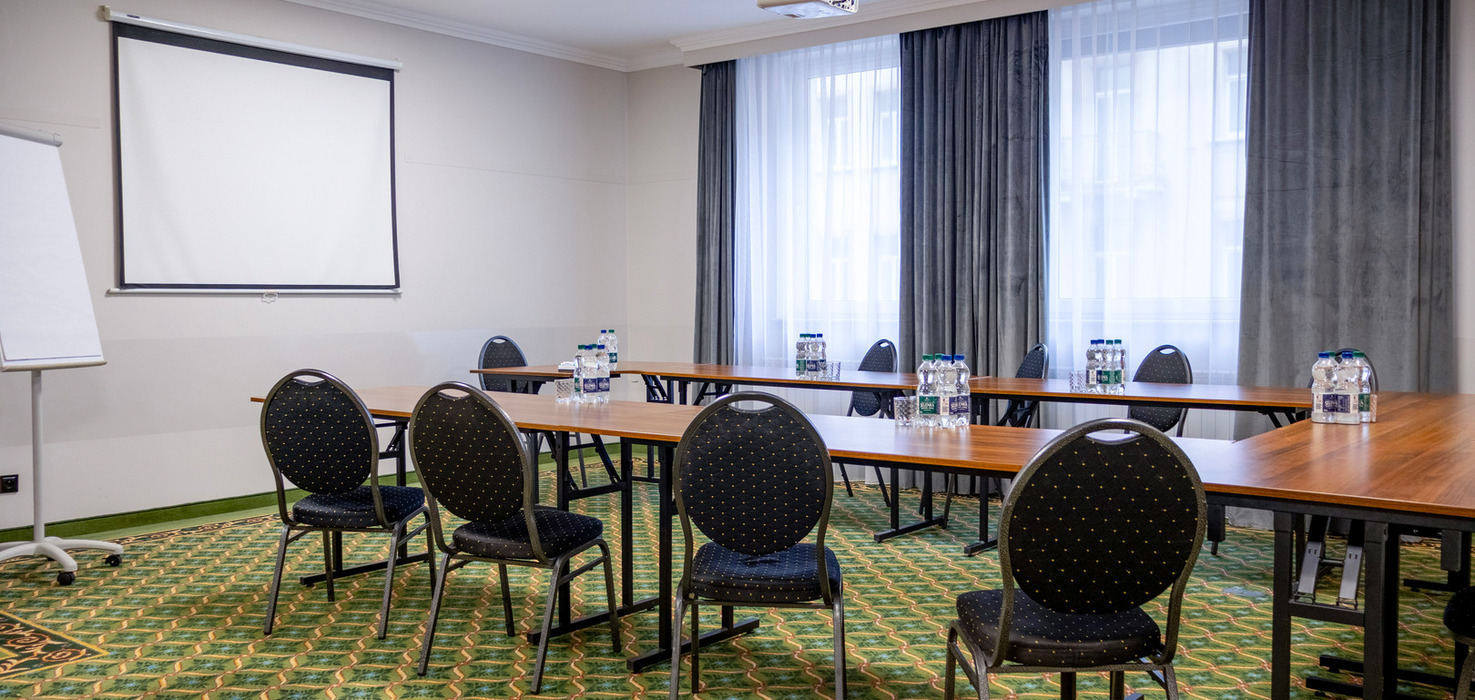 Sala konferencyjna hotelu w układzie U, projektor sufitowy i ekran, butelki wody, wzorzysty dywan, firany