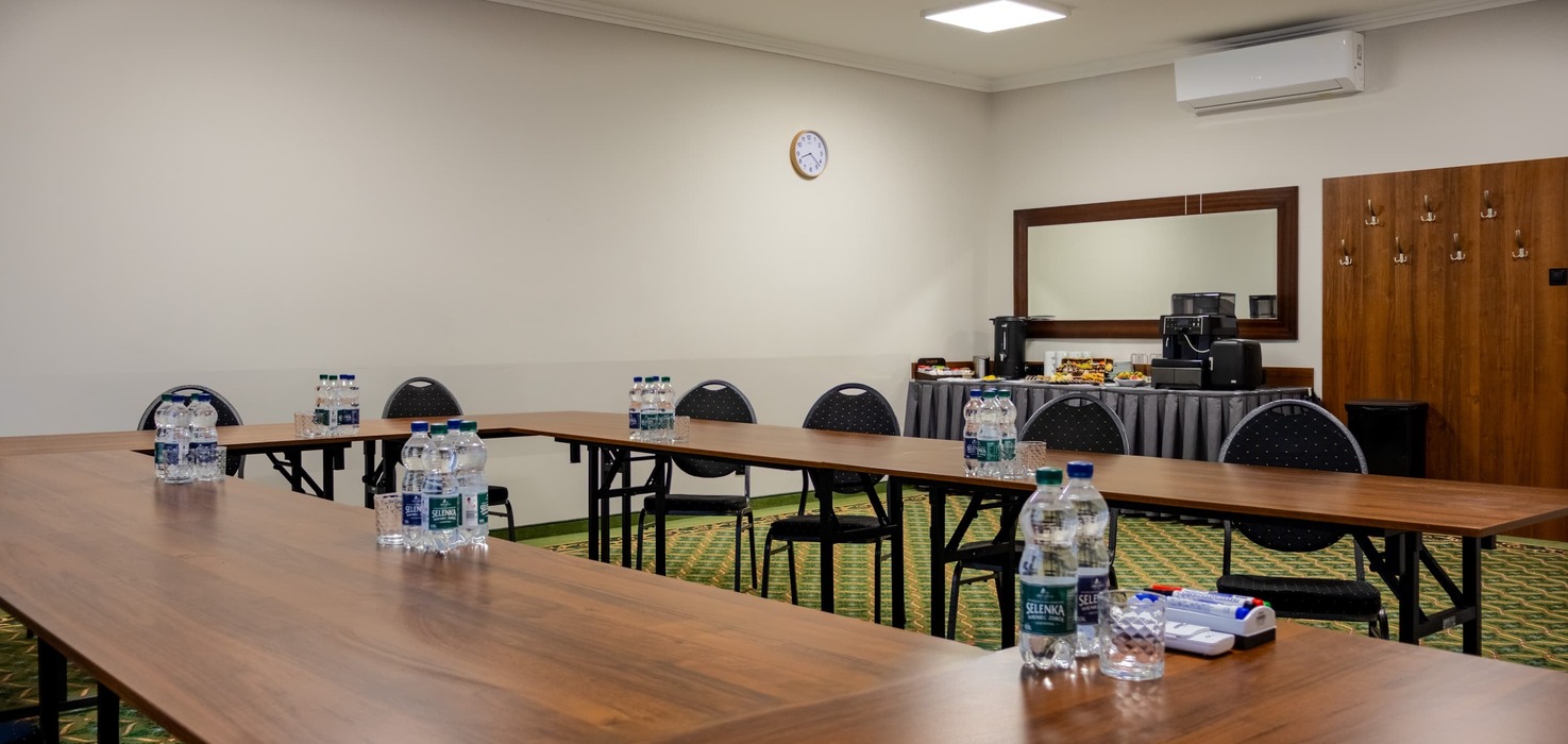 Conference room with tables arranged in a horseshoe shape, with water bottles prepared on them. In the background, there is a coffee service area and a large wall mirror, and a projector is suspended from the ceiling.