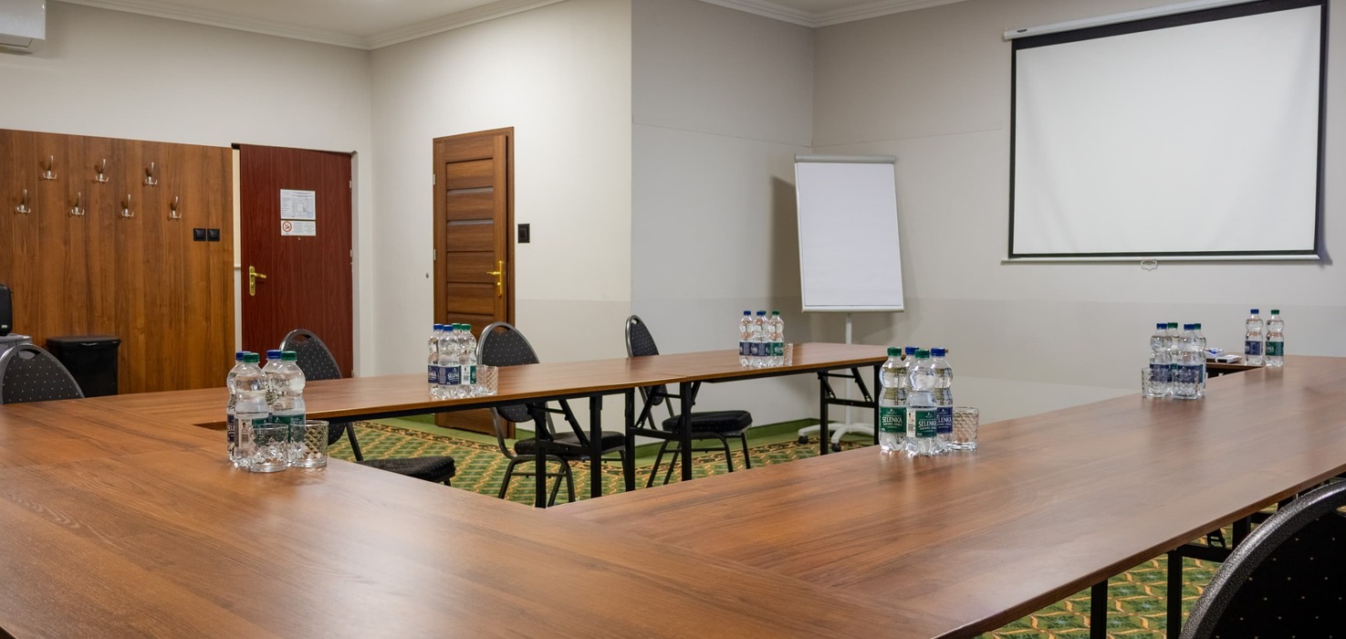 Conference room with tables arranged in a horseshoe shape, with water bottles prepared for participants on them. In the background, a projector suspended from the ceiling, a projection screen, a flipchart, and two wooden doors are visible.