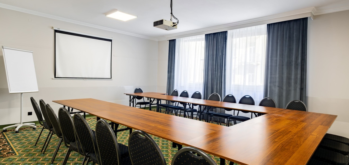 Conference room with tables arranged in a horseshoe layout, a projector suspended from the ceiling, a projection screen on the side wall, and windows with dark curtains.