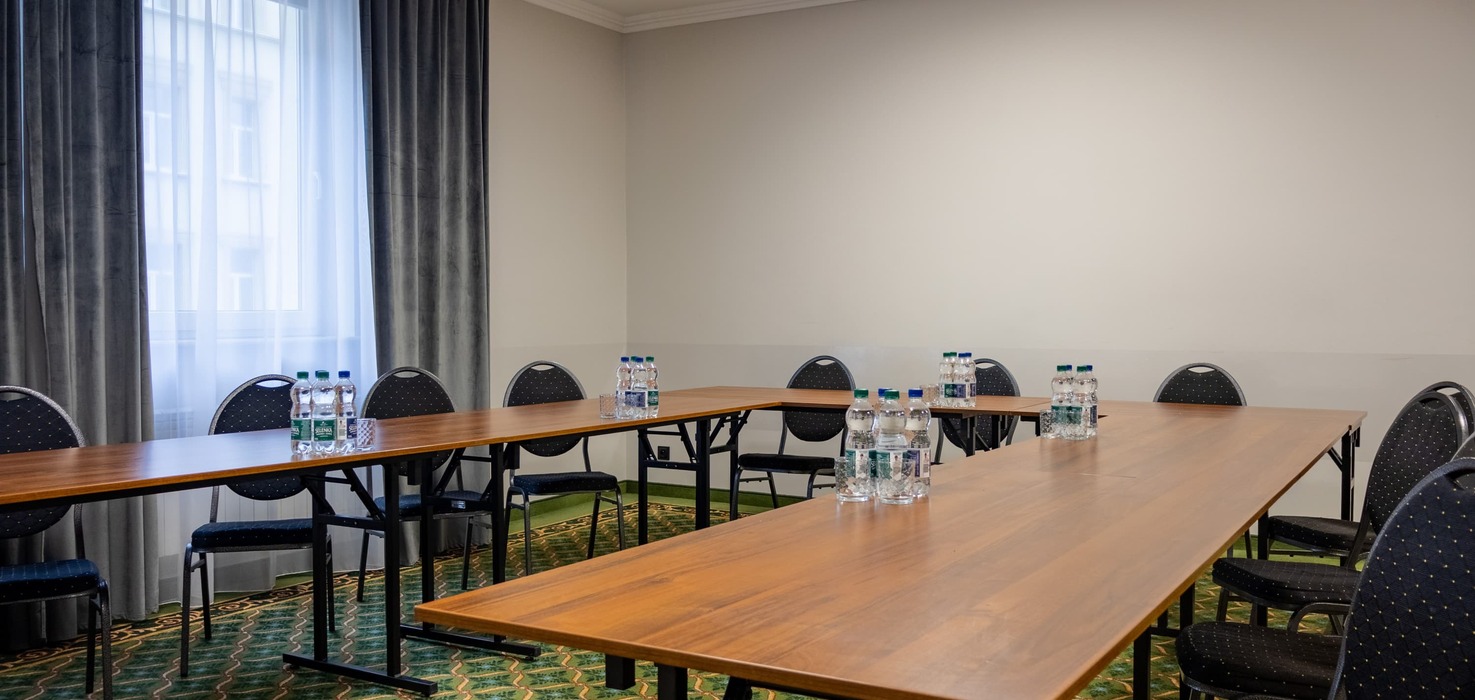 Conference room with tables arranged in a horseshoe layout, with water bottles prepared for participants on them. A projector is mounted under the ceiling, and on the left side there is a window with dark curtains.