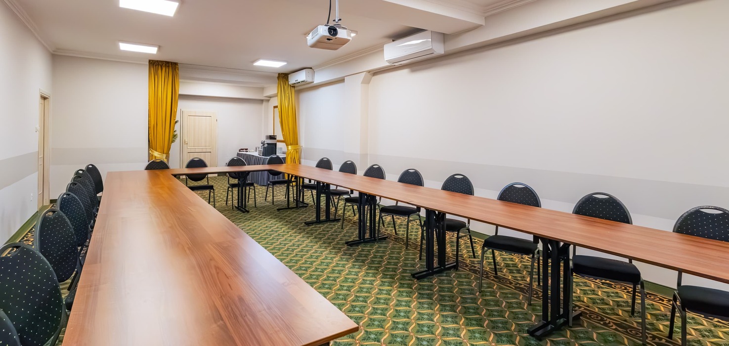 Conference room with tables arranged in a horseshoe layout, a projector suspended from the ceiling, and a visible coffee service area in the background.