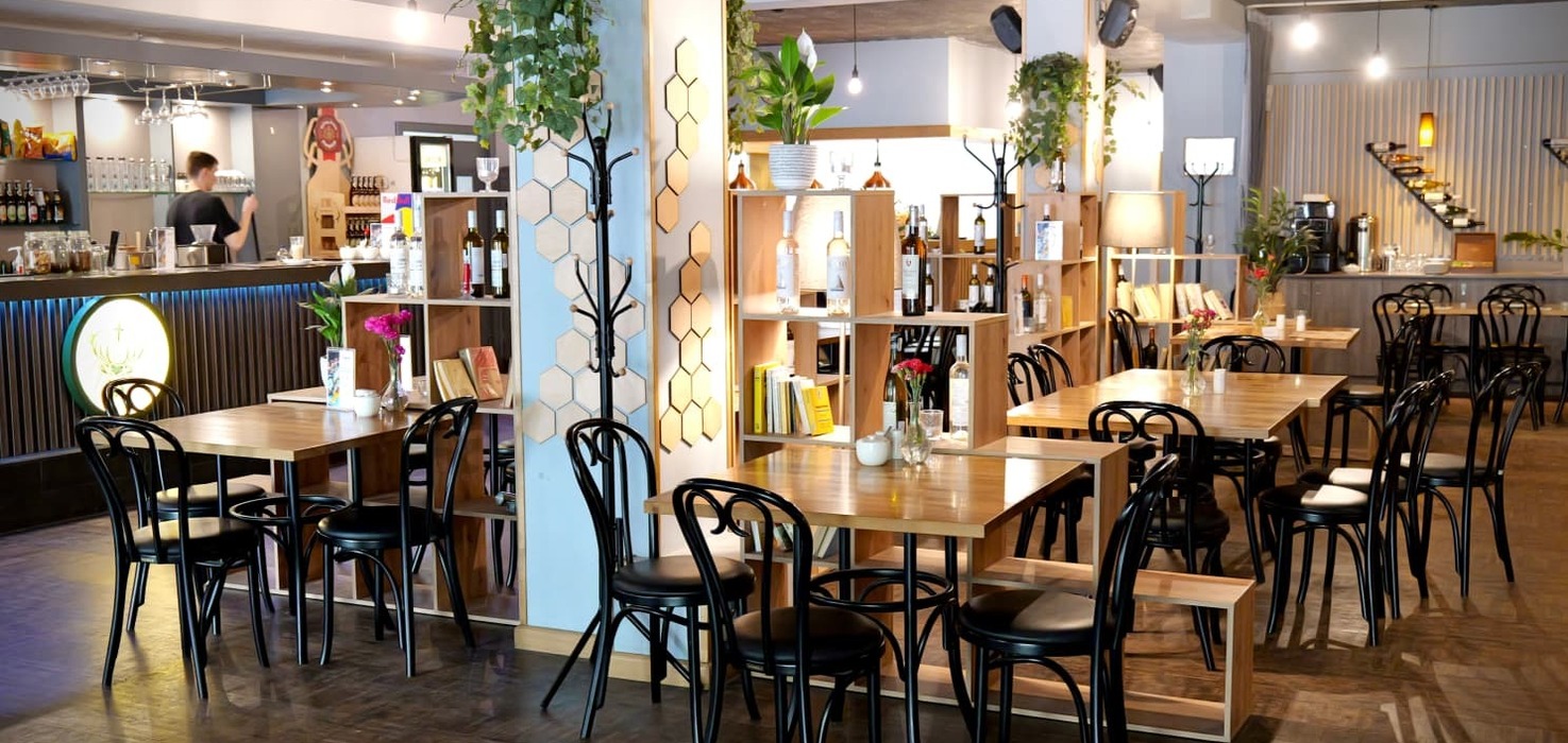 Modern hotel café with wooden tables, black chairs, open shelves of wines and books, hanging plants, pendant lights and a staff member at the bar.