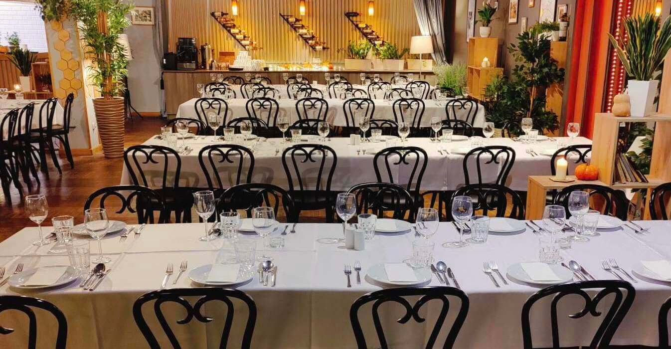Banquet dining room with long white‑clothed tables, elegant place settings, black curved chairs and warm ambient lighting.