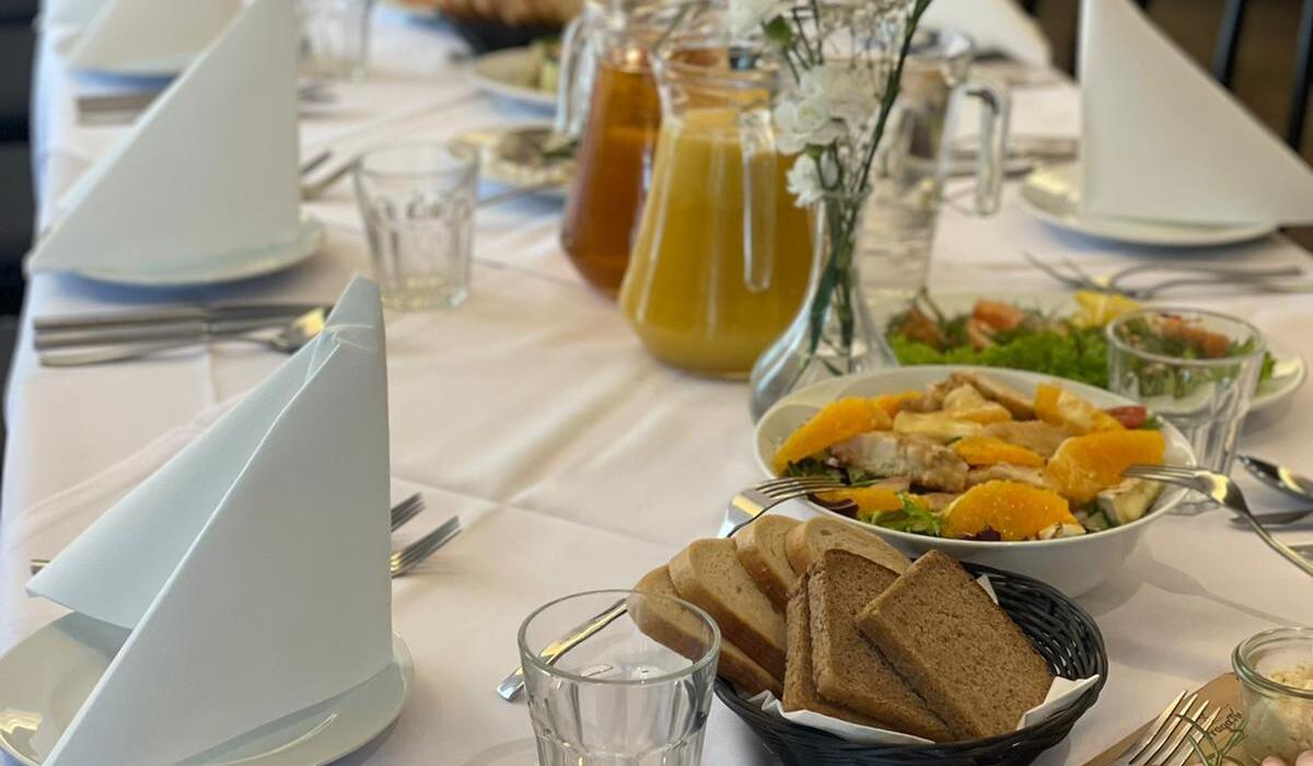 Lange Banketttafel mit weißen Servietten, Gläsern, Saftkannen, Salaten, Brotkörben, Aufschnitt und kleinen weißen Blumen