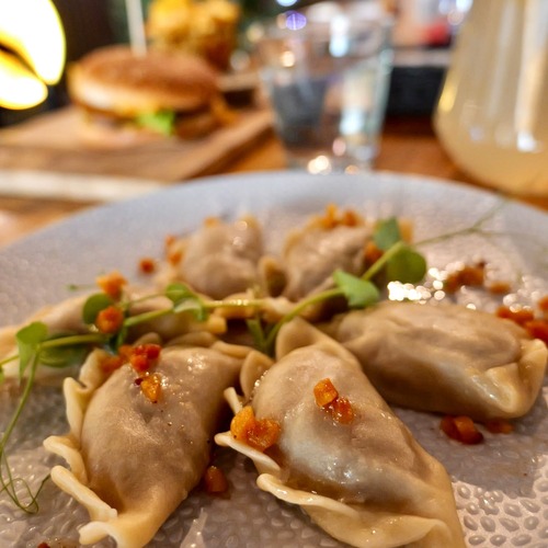 Talerz domowych pierogów z chrupiącymi dodatkami i mikro ziołami, w tle rozmyty burger i orzeźwiający napój – domowe smaki i wyjątkowa oferta restauracji PragaTu w Warszawie blisko stacji metra Dworzec Wileński.