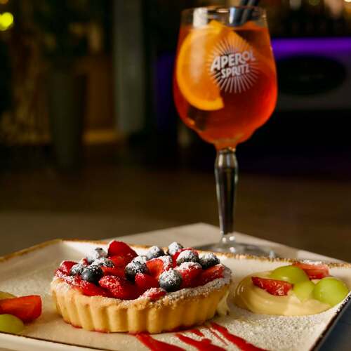 Kusi Cię słodka chwila relaksacja? Pyszna tarta z truskawkami i jagodami posypana cukrem pudrem, podana z kremem, winogronami i orzeźwiającym Aperol Spritz – idealny deser w klimatycznym barze restauracji PragaTu w Warszawie blisko stacji metra Dworzec Wileński.