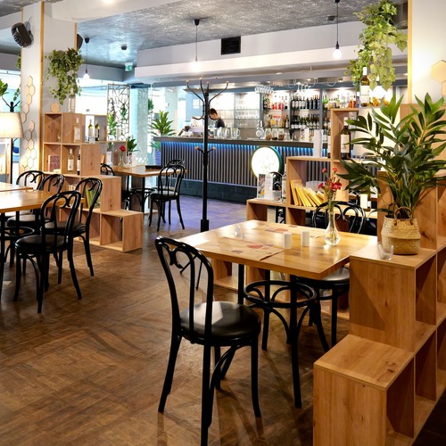 Nowoczesna restauracja hotelowa z eleganckimi drewnianymi stolikami, czarnymi krzesłami, bujnymi roślinami, stylowymi wiszącymi lampami i klimatycznym barkiem – przytulna przestrzeń i doskonałe smaki w restauracji PragaTu w Warszawie blisko stacji metra Dworzec Wileński.