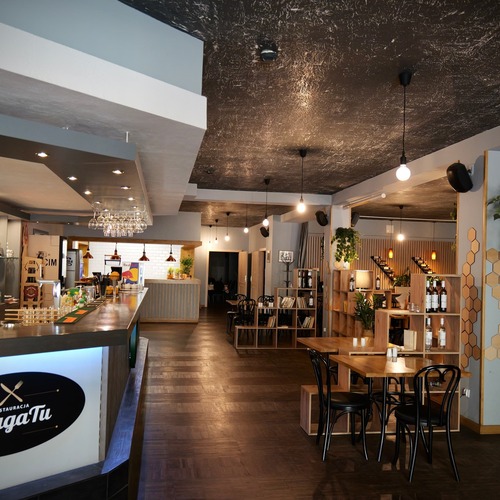 Nowoczesna restauracja z klimatycznym barem w hotelu, eleganckimi drewnianymi stołami, regałami z wyselekcjonowanymi winami, stylowymi wiszącymi lampami i przytulną, otwartą jadalnią – idealne miejsce na spotkania i doskonałe smaki w restauracji PragaTu w Warszawie blisko stacji metra Dworzec Wileński.