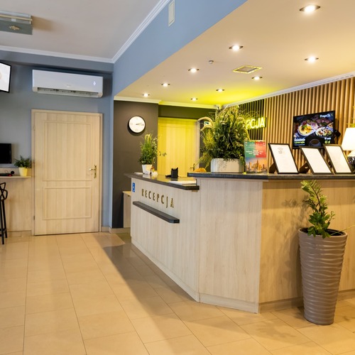 Nowoczesna recepcja hotelowa z eleganckim drewnianym biurkiem, ciepłym nastrojowym oświetleniem, bujnymi roślinami, czytelnymi broszurami i nowoczesnym ekranem informacyjnym – profesjonalna obsługa i przyjazna atmosfera w Hit Hotel w Warszawie blisko stacji metra Dworzec Wileński.