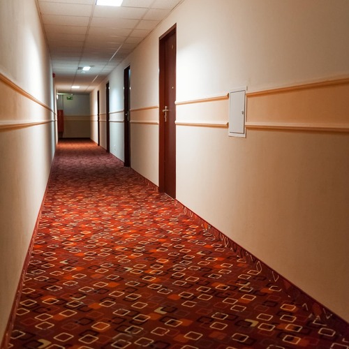 Długi hotelowy korytarz z eleganckim czerwonym wzorzystym dywanem, kremowymi ścianami, stylowymi drzwiami do pokoi i rzędem sufitowych lamp prowadzących w kierunku wyjścia – komfortowy wypoczynek i przytulna atmosfera w Hit Hotel w Warszawie blisko stacji metra Dworzec Wileński.