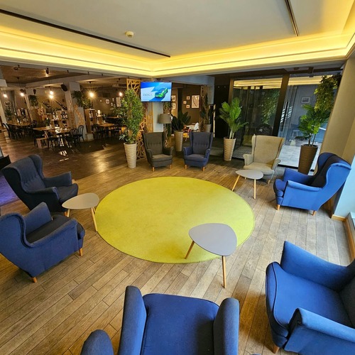 Przytulny hotelowy salon z eleganckimi niebieskimi fotelami rozstawionymi wokół dużego, słonecznego żółtego dywanu, stylowymi stolikami, bujnymi roślinami i ciepłym oświetleniem tuż obok części restauracyjnej – idealne miejsce na relaks i doskonałe smaki w Hit Hotel w Warszawie blisko stacji metra Dworzec Wileński.