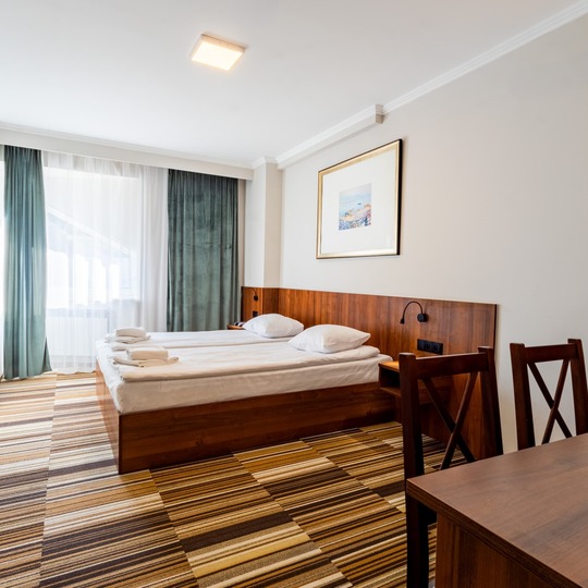Bright hotel room with two single beds, wooden headboard, bedside reading lights, wall TV, sheer and green curtains, desk and striped carpet.
