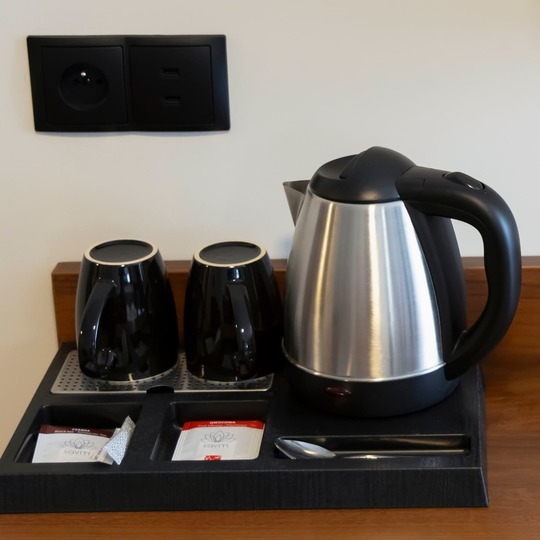 Electric kettle and black cups on a tray beside a modern lamp with a power socket. A hot drink in the room is a great convenience. By booking accommodation in Warsaw, you gain complete comfort.