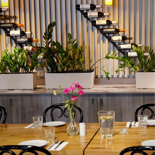 Przytulny stół restauracyjny z drewnianym blatem, wazonem z różowymi kwiatami, dzbankiem wody i szklankami; lamelowana ściana, donice i półki na wino.