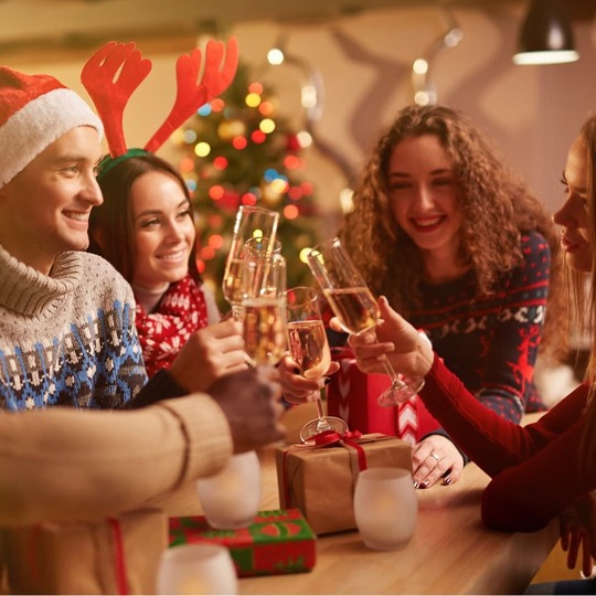 Guests in festive sweaters and Santa hats toast with champagne around a gift-strewn table by a decorated Christmas tree.