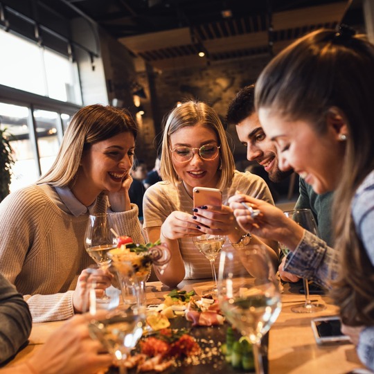 Friends laughing over a phone while sharing small plates and wine at a cozy modern restaurant with warm lighting and large windows