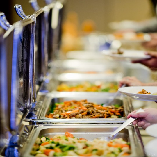 Hotel buffet line with chafing dishes and guests serving plated hot dishes in a warm-lit dining area
