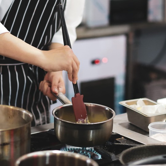 Chef in striped apron stirring sauce on a gas stove in a professional kitchen, pans and croquettes nearby