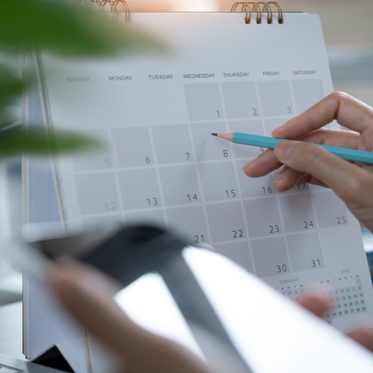 Hand holding a pencil marking a date on a desk calendar beside a blurred smartphone and plant
