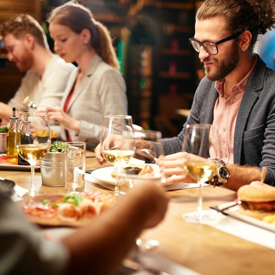 Group enjoying a casual dinner in the hotel restaurant — communal wooden table, wine glasses, gourmet burger, salads and warm lighting.