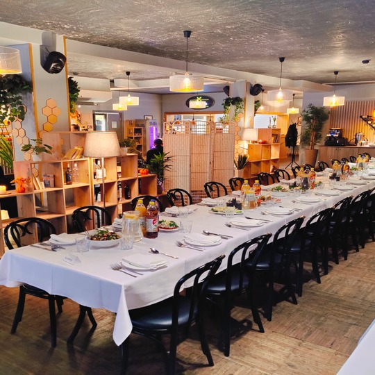 Long banquet table with white linen, plates and salads in a cozy modern restaurant with pendant lights and bar
