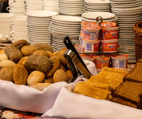 Breakfast buffet with assorted bread rolls and sliced loaves, stacked white plates, yogurt cups and a fruit basket
