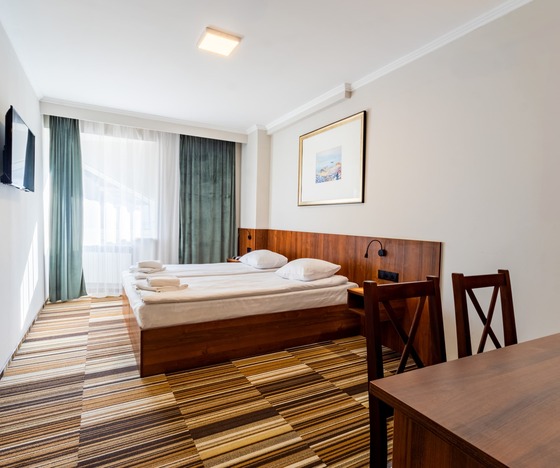 Helles Hotelzimmer mit zwei Einzelbetten, Holzkopfteil, Leseleuchten, Wand-TV, transparenten und grünen Vorhängen, Schreibtisch und gestreiftem Teppich.