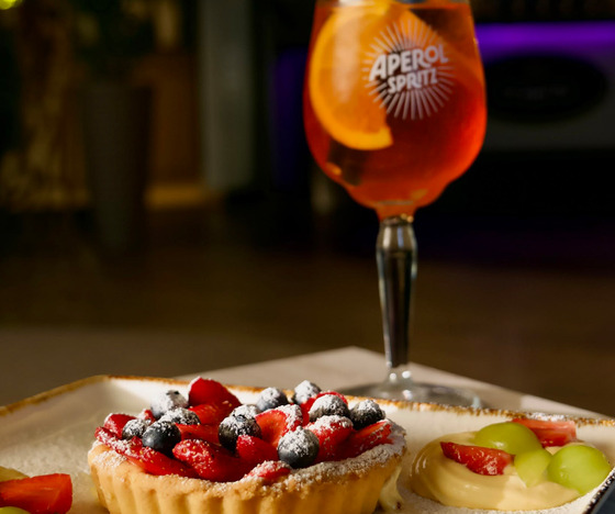 Tarta z jagodami oprószona cukrem pudrem obok kremowej tarty z owocami, z Aperol Spritz i plastrem pomarańczy przy barze.