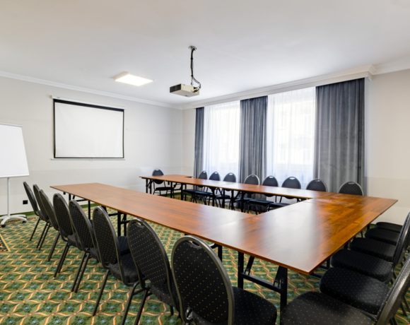 Sala konferencyjna z układem stołów w kształcie litery U, krzesła, ekran do projektora i flipchart, jasne wnętrze z dużymi oknami.