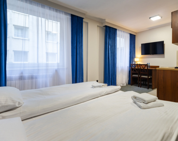 Pokój hotelowy z dwoma pojedynczymi łóżkami, niebieskimi zasłonami, biurkiem, telewizorem i małym aneksem kuchennym.