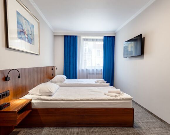 Hotel room with two single beds, white bedding, blue curtains, and a wall-mounted TV