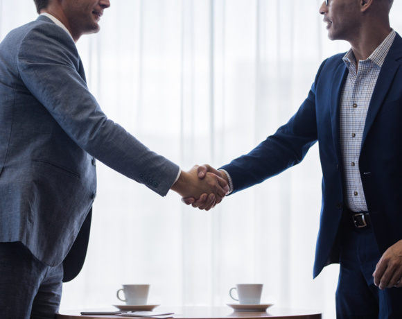 Two men in suits shake hands over a table with two coffee cups, in a professional meeting setting