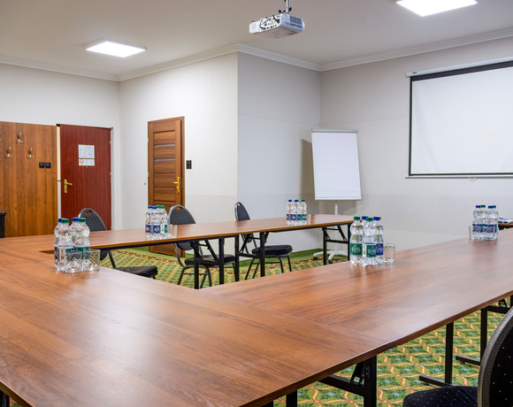 Sala konferencyjna w układzie U z projektorem, ekranem, flipchartem i butelkowaną wodą na ozdobnym dywanie.
