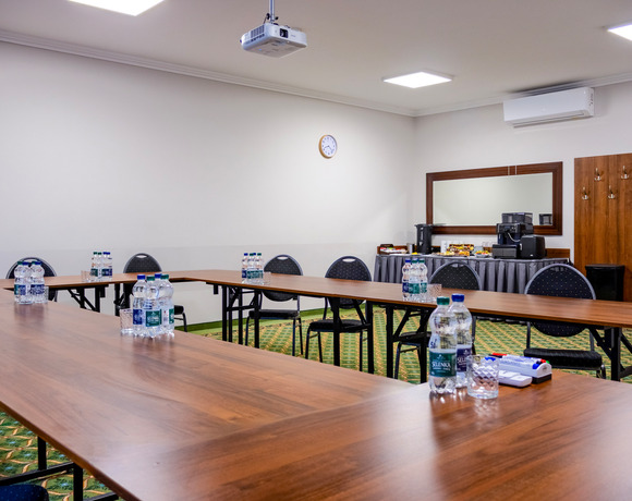 Sala konferencyjna w układzie U z drewnianymi stołami, butelkowaną wodą, projektorem sufitowym, stacją kawową i klimatyzacją.