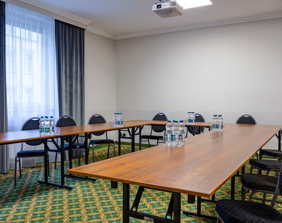 Mała sala konferencyjna hotelu z stołami w układzie U, czarnymi krzesłami, butelkami wody, projektorem i zasłoniętym oknem