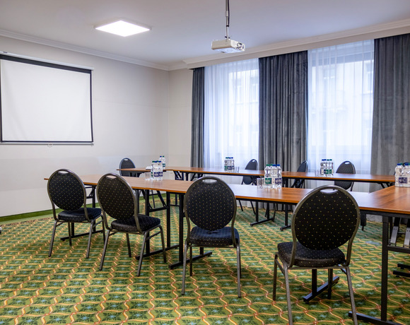 Sala konferencyjna hotelu w układzie U, projektor sufitowy i ekran, butelki wody, wzorzysty dywan, firany