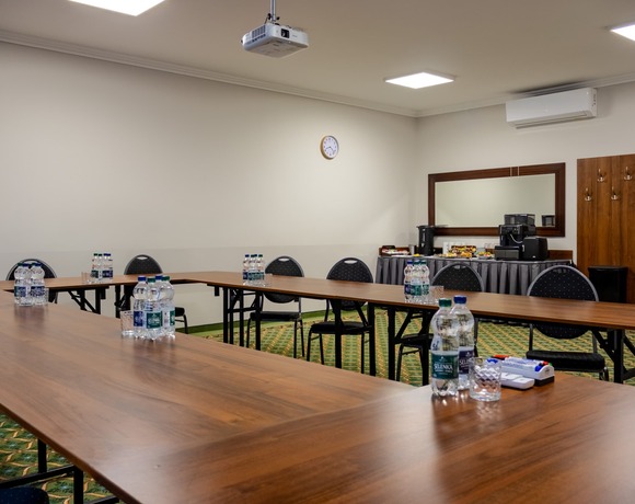 Conference room with tables arranged in a horseshoe shape, with water bottles prepared on them. In the background, there is a coffee service area and a large wall mirror, and a projector is suspended from the ceiling.