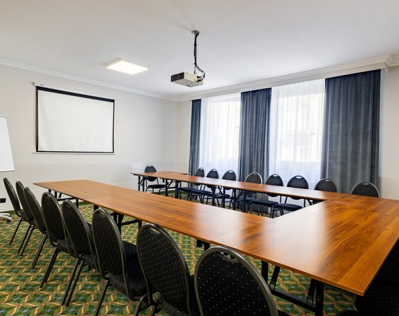 Conference room with tables arranged in a horseshoe layout, a projector suspended from the ceiling, a projection screen on the side wall, and windows with dark curtains.