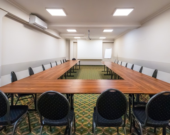 Klimatyzowana sala konferencyjna z układem stołów w kształcie litery U, czarnymi tapicerowanymi krzesłami, projektorem, ekranem i flipchartem – profesjonalna przestrzeń biznesowa w obiekcie oferującym hotel Warszawa blisko stacji metra Dworzec Wileński.